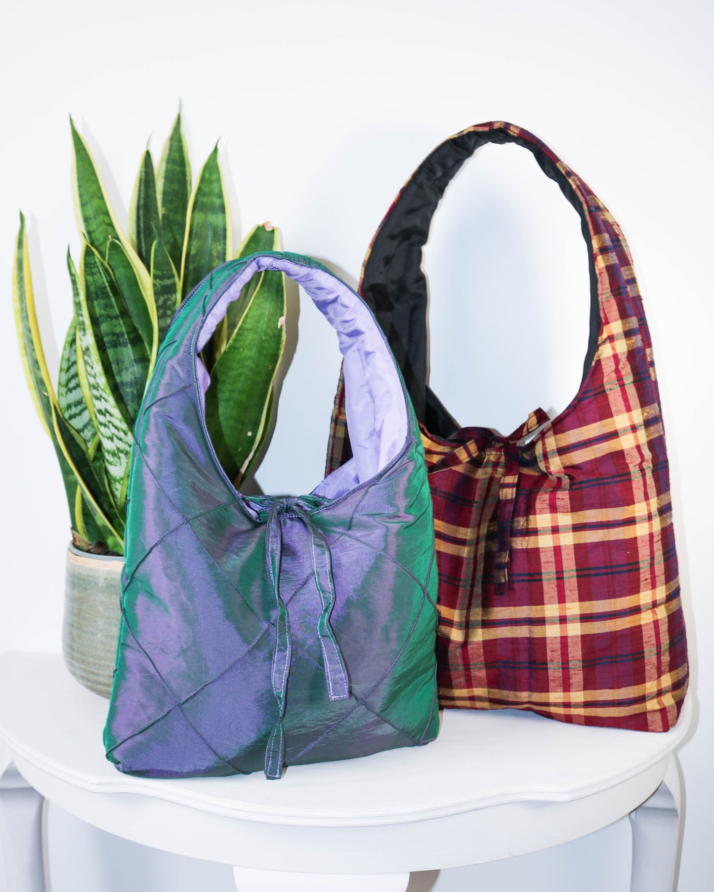 Two handbags, one green and one plaid, on a white surface with a plant in the background.